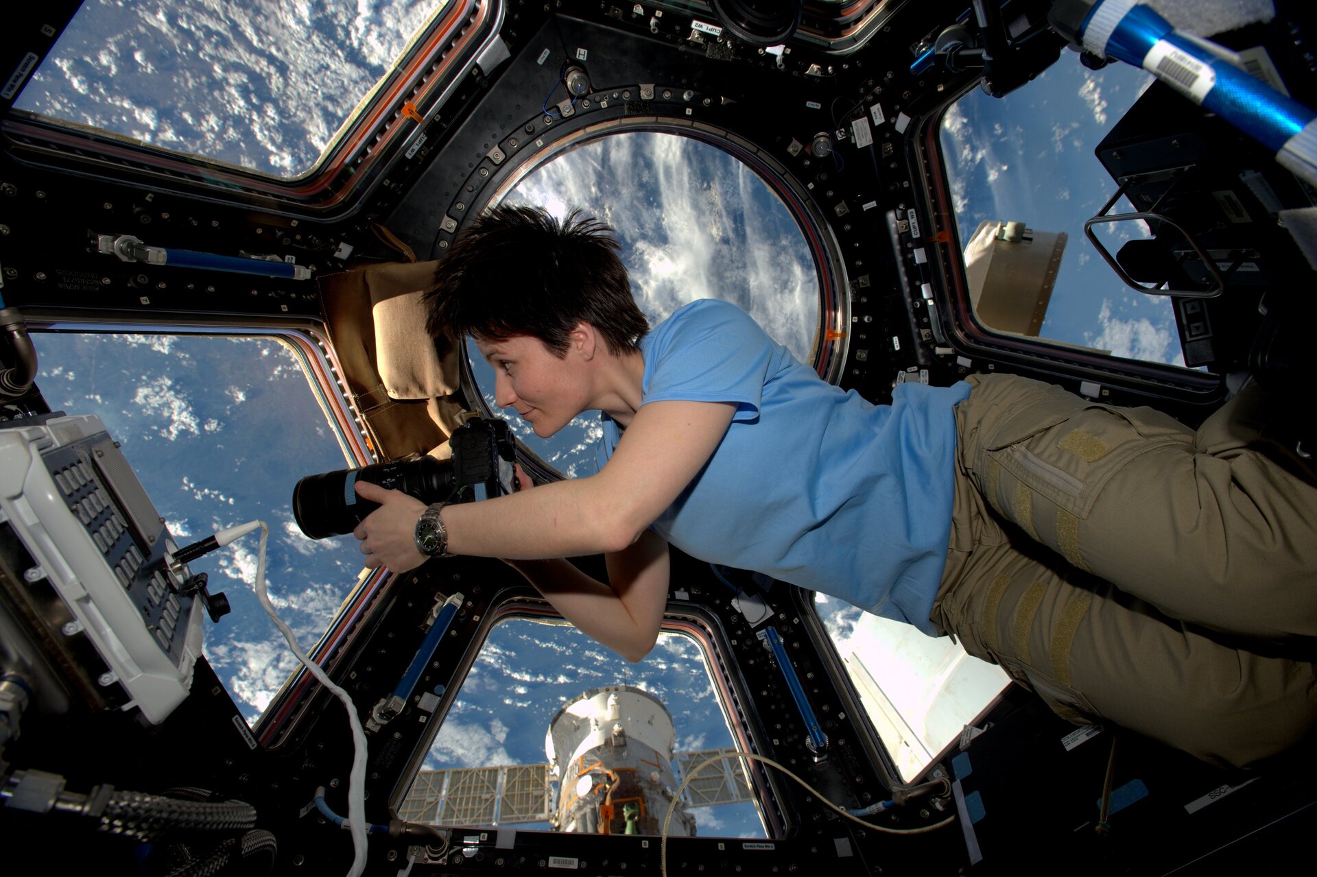 Die Astronautin Samantha Cristoforetti schwerelos auf der ISS mit einer Kamera in ihren Händen. Ausserhalb des Fensters kann man die Erdoberfläche sehen.